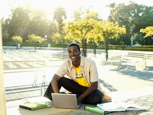 Male university student sitting on ground with textbooks, using laptop, smiling, portrait