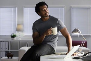 Serious Black man sitting on desk in home office drinking coffee